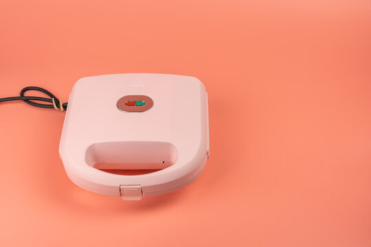 A closed pink sandwich maker with a red and green power indicator light sits on a solid coral background with open copy space.