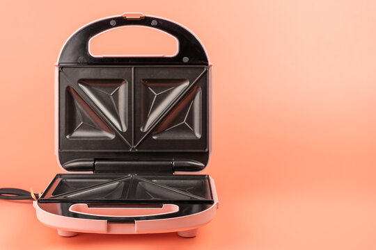 An open pink sandwich maker faces forward, showing its symmetrical black triangle plates and handle on a solid coral background.