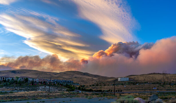Smoke billowing from a destructive wildfire in the Peavine Mountains, north of Reno, Nevada at sunset The sky is has vibrant pink and orange clouds of smoke contrasting with the blue sky. 