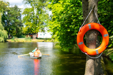 Orange lifebuoy ring hanging on a tree trunk in the foreground, representing water safety, with a blurred background of a rowing boat on a park lake in bright summer sunlight. © Adam