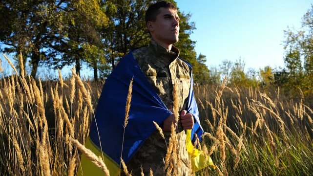 Military man in uniform stands with flag of Ukraine on shoulders at countryside. Portrait of male ukrainian soldier with national banner in honor of victory against russian aggression. End of war