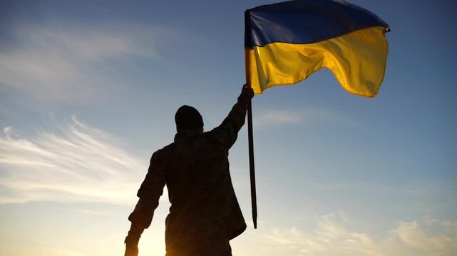 Silhouette of male military lifting blue-yellow banner in honor of the victory against russian aggression at sunset. Soldier of ukrainian army raised a waving flag of Ukraine at countryside. Slow mo