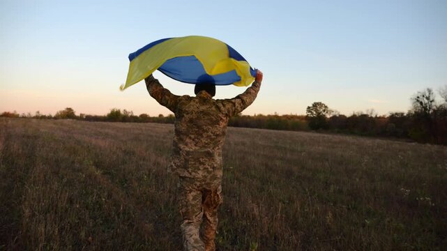 Soldier of ukrainian army running with raised flag Ukraine above his head at field. Young male military in uniform jogging with blue-yellow banner at meadow. Victory against russian aggression
