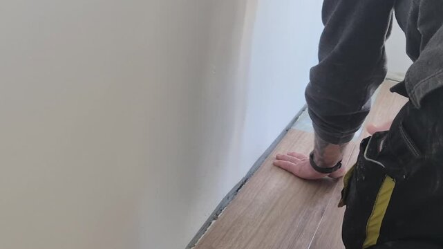 Professional installer laying vinyl flooring along the wall precisely, smoothing it with his hand in a new apartment.