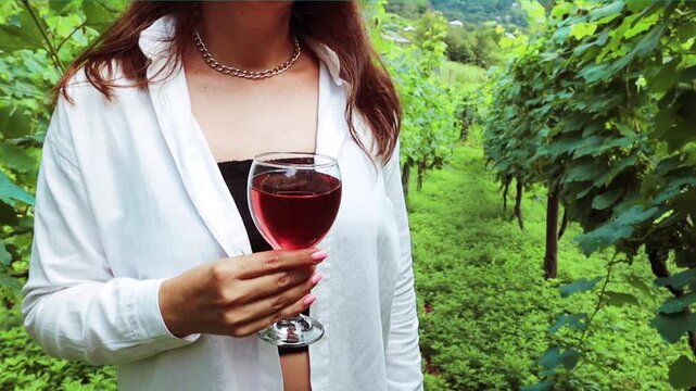 Woman unrecognized wearing the white shirt holding the glass of rose wine standing at the green vinery farn in Georgia