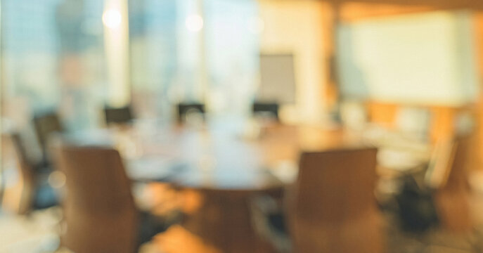 A blurred image of a modern conference room with a round table surrounded by chairs. Large windows allow natural light to fill the space, enhancing the professional atmosphere.