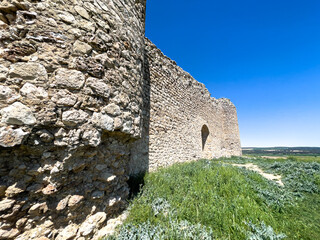 A weathered medieval stone castle wall with a round tower rising above green grassy fields, set against a vibrant clear blue sky on a sunny day. © Licia.Galli