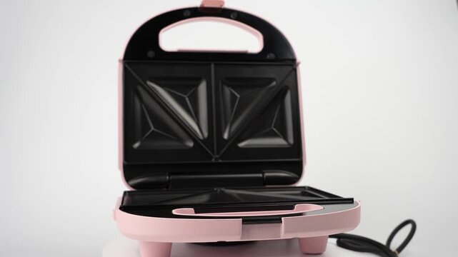 A pink and black sandwich maker sits open on a white studio background, rotating gently to reveal its non-stick plates, feet, and coiled cord.