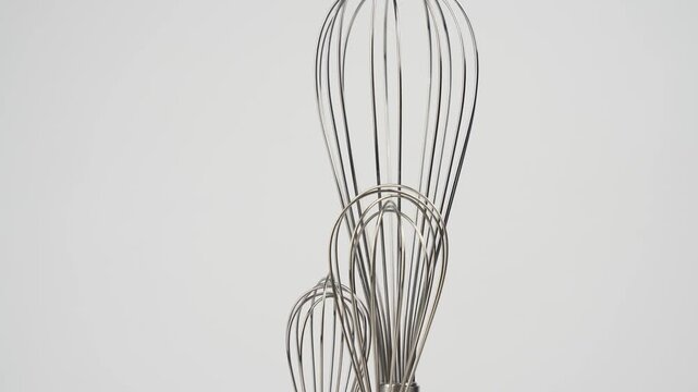 Three metal whisks of different sizes stand bundled together against a plain white background in a close, tightly framed studio shot.