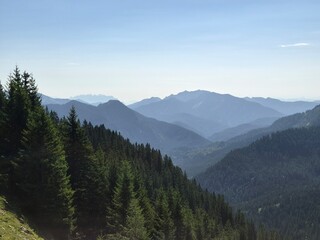 Fototapeta premium Landscape of mountain with a clear sky above a deep forested valley and distant, layered blue Alpine peaks in soft haze. Bird Flying View at Deep Alpine Valley and Mountain Peaks