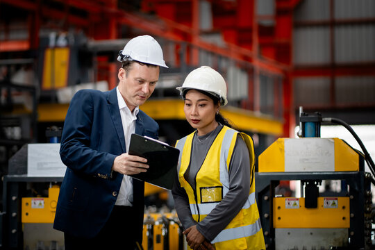 Two professional engineer,worker,technician use clipboard discuss work, walk in steel metal manufacture factory plant industry. Black African American man and woman wear hard hat check quality machine