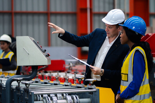 Two professional engineer,worker,technician use clipboard discuss work, walk in steel metal manufacture factory plant industry. Black African American man and woman wear hard hat check quality machine