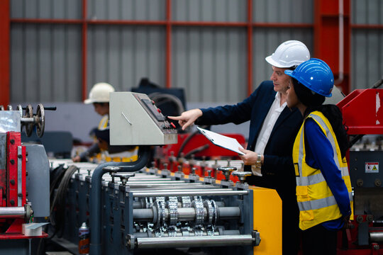 Two professional engineer,worker,technician use clipboard discuss work, walk in steel metal manufacture factory plant industry. Black African American man and woman wear hard hat check quality machine