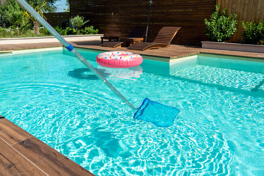 Skimmer net cleaning turquoise swimming pool water with wooden deck and colorful inflatable ring
