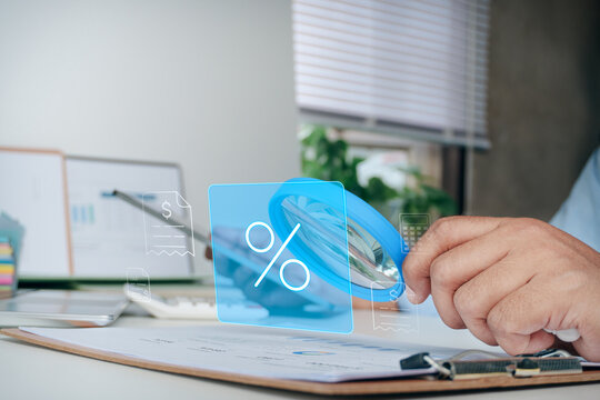 Hand holding magnifying glass focusing on percentage interest rate and financial document hologram icons, concept of tax calculation, mortgage loan interest, business investment, and banking finance.