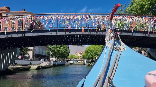 POV shot from the prow of a traditional Moliceiro boat passing under a bridge in Aveiro, Portugal, as another colorful tourist boat approaches on a sunny day.