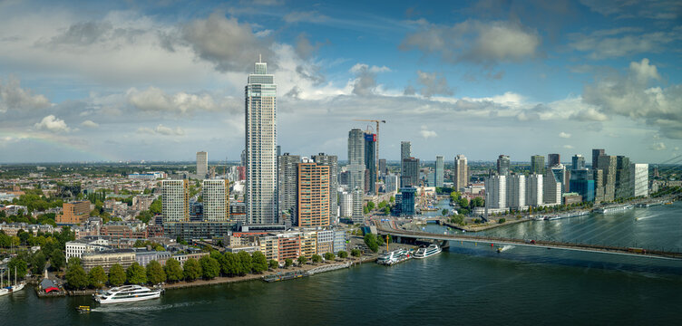 Panoramic view of Rotterdam skyline and the Nieuwe Maas river under dramatic clouds and blue sky, featuring modern high-rise architecture.