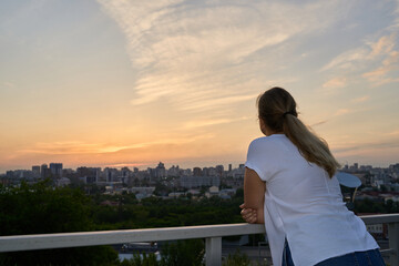 Fototapeta premium Young Woman Leaning On Balcony Railing Watching Distant City Skyline Under Warm Sunset Clouds From Elevated Rooftop Terrace.