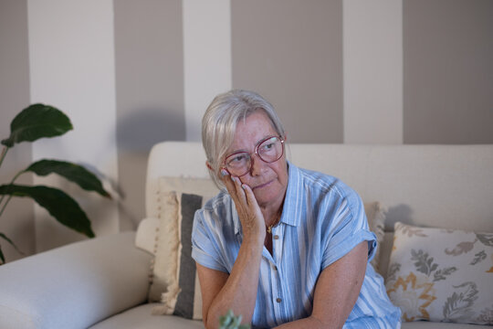 Senior adult woman feeling lonely and sad at home, sitting on a sofa in the living room, experiencing depression, looking pensive and thoughtful, needing care and emotional support