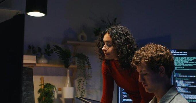 A Latin engineer uses a digital tablet to explain and correct server data as teammates at computers collaborate, reviewing database entries, network logs, and security settings.