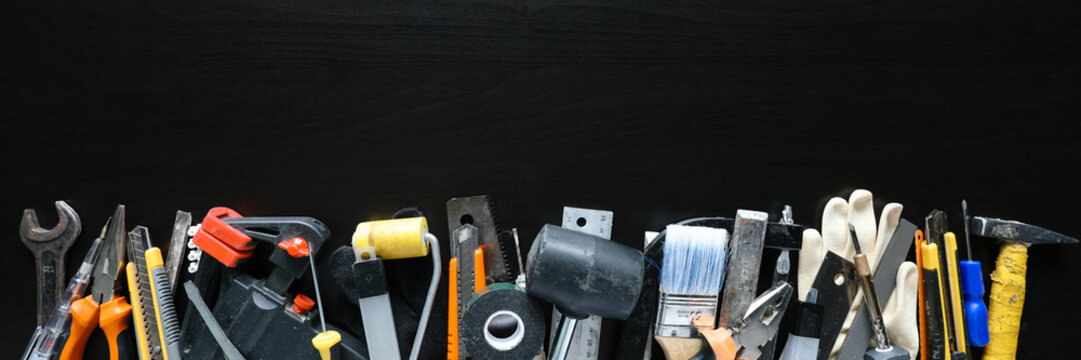 Assorted hand tools, paint brush, gloves, utility knives, pliers, wrench, tape, rulers and hammer arranged in row on black background for repair