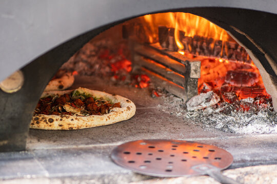 Pizza baking in a wood-fired stone pizza oven with a roaring log fire in the back