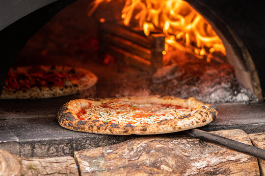 Pizzas baking in a wood-fired stone pizza oven with a roaring log fire in the back