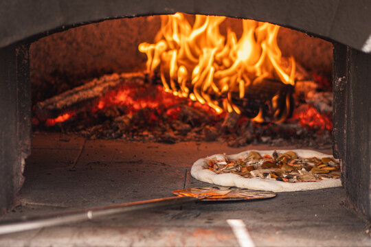 Pizza baking in a wood-fired stone pizza oven with a roaring log fire in the back