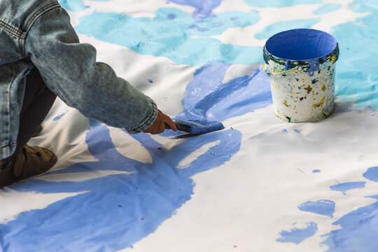 Child using a paintbrush to spread vibrant blue paint over a large white surface next to an open bucket of matching blue paint