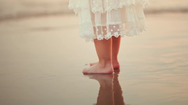 Close up of barefoot little girl walking through shallow seawater on sandy beach, child feet splashing gentle waves while wearing white dress, peaceful summer vacation and childhood outdoor lifestyle