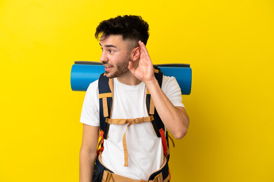 Young mountaineer caucasian man with a big backpack isolated on yellow background listening to something by putting hand on the ear