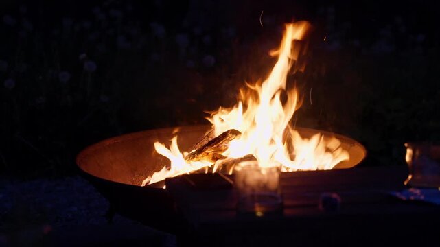 Bright flames rise from stacked wood inside outdoor metal fire pit, with warm orange glow, deep shadow, soft foreground blur, and intimate cozy atmosphere in low light outdoor setting