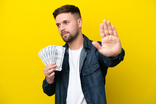 Young caucasian man taking a lot of money  isolated on yellow background making stop gesture and disappointed