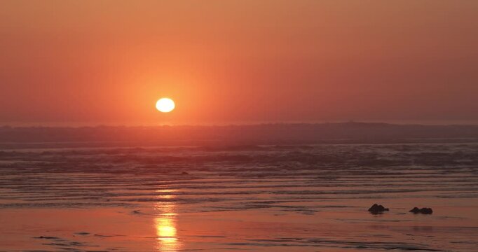 rolling ocean sea waves crash gently on a serene beach at sunset orange skies and silver blue surf filmed during golden hour in Cornwall, UK with small rocks as the sun sets over the horizon 4K video
