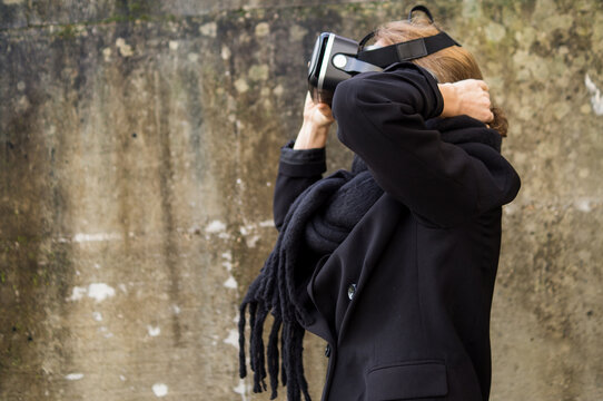 Medium shot of a stylish woman wearing a black winter coat and scarf adjusting a VR headset over her eyes with a concrete wall background.