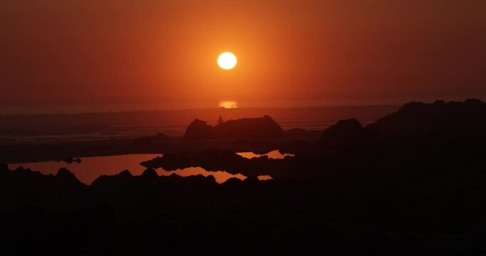 stunning exotic sunset of a brilliant orange glowing sun slowly setting over the horizon of the ocean with a rocky silhouette beach shoreline and rockpools in the foreground with some joggers running 