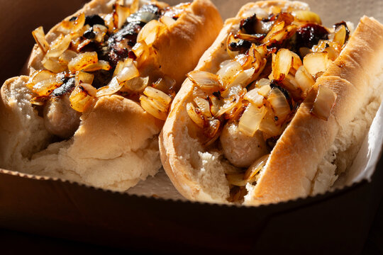 Hot dogs with fried onions in white finger bread rolls, made with pork sausages, in a cardboard takeaway tray background backdrop.