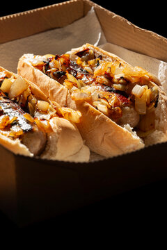 Hot dogs with fried onions in white finger bread rolls, made with pork sausages, in a cardboard takeaway tray background backdrop.