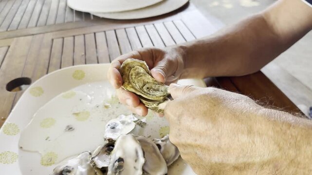 Close-up of two male hands opening a fresh oyster from the rear hinge using an oyster knife. Traditional preparation of raw shellfish before serving, with an oyster from Cap Fer