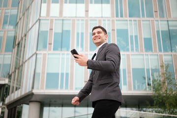 Fototapeta premium Businessman smiles while walking and checking phone outside modern office building