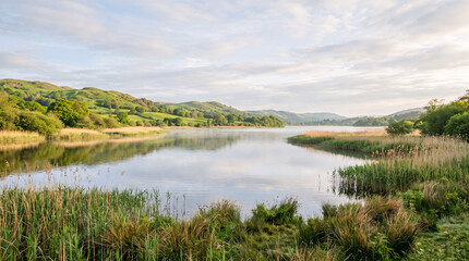 Fototapeta premium Peaceful landscape of a tranquil lake reflecting the cloudy sky with green hills in the background.