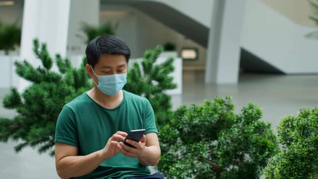 Young adult man wearing surgical mask seated in bright indoor atrium, tapping smartphone with both hands while looking down in focused posture beside modern plants and architecture