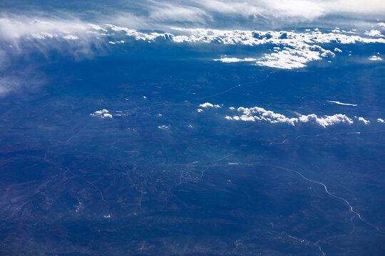 Airplane window view captures dramatic wispy clouds stretching across the earth. Scattered cumulus clouds cast soft dark shadows over the vast blue valleys and rolling mountain terrain far below