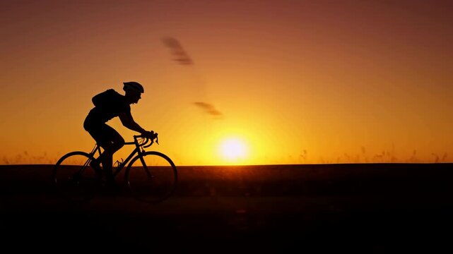 Cyclist riding along open landscape in silhouette during golden sunset with bicycle wheels moving across horizon line, warm orange sky, tall grasses, and dramatic backlit outdoor scenery