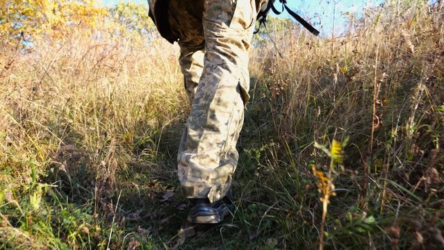 Low view to male feet of ukrainian soldiers going along trail at countryside. Legs of young army man walking through dry grass at sunny day. War in Ukraine. Slow motion