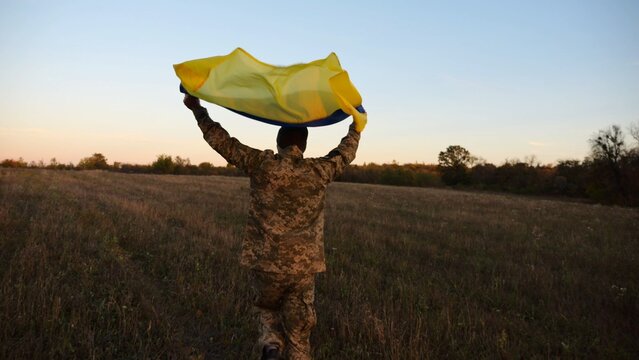 Soldier of ukrainian army running with raised flag Ukraine above his head at field. Young male military in uniform jogging with blue-yellow banner at meadow. Victory against russian aggression
