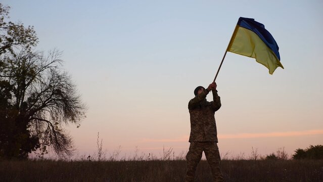 Male military waving blue-yellow banner on liberated territory occupied by russia. Young soldier of ukrainian army lifted flag of Ukraine in honor of victory against russian aggression at dusk