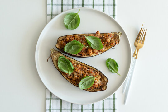 Two halves of baked eggplant stuffed with savory meat and vegetable filling garnished with fresh basil leaves served on a white plate with golden forks beside it.