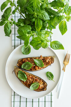 Two stuffed eggplant halves presented on white plate with fresh basil garnish and golden cutlery on checkered napkin