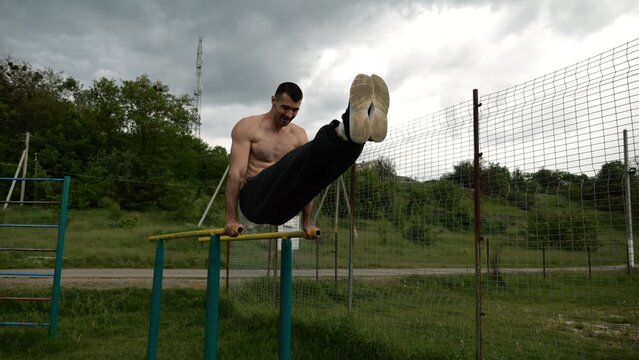 Strong man trains abdominal muscles raising legs on parallel bars at sports ground. Young shirtless sportsman exercising at nature. Sporty athlete working out at nature. Concept of sportive lifestyle